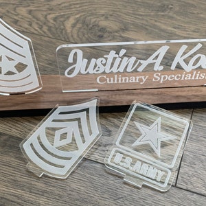May include: A wooden plaque with a clear acrylic nameplate that reads "Justin A Koons Culinary Specialist". There are three clear acrylic military insignia: a star, a chevron, and a chevron with a star.