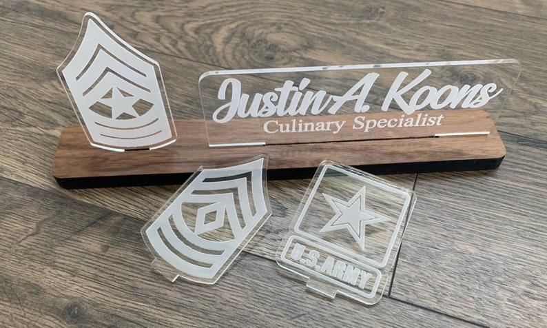 May include: A wooden plaque with a clear acrylic nameplate that reads "Justin A Koons Culinary Specialist". There are three clear acrylic military insignia: a star, a chevron, and a chevron with a star.
