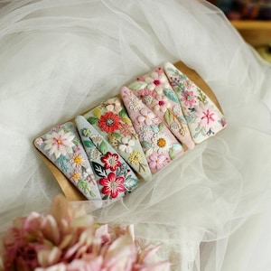 Könnte beinhalten: Sechs bestickte Haarspangen mit floralen Designs in verschiedenen Farben, darunter Rosa, Rot, Weiß und Grün. Die Spangen sind auf einer braunen Karte angeordnet und zeigen detaillierte Stickereien. Jede Spange hat ein anderes Blumenarrangement.