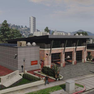 Op de afbeelding: Buitenaanzicht van het San Andreas Fire Department-gebouw. Het gebouw heeft een modern ontwerp met een combinatie van baksteen en grijs beton. Een Amerikaanse vlag wappert op de voorgrond. Het gebouw heeft meerdere garagedeuren.