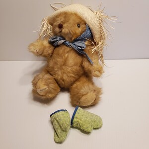 May include: A brown teddy bear wearing a straw hat with a blue and white bandana around its neck. The bear is sitting on a white surface and has green and blue mittens next to it.