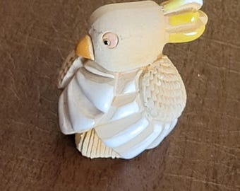 May include: A small, white ceramic bird figurine with a yellow crest and a detailed, textured body. The bird is perched on a wooden surface.