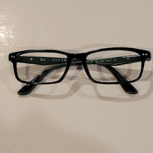May include: Black rectangular eyeglasses with clear lenses. The glasses have a thick frame and the Ray-Ban logo is visible on the left arm. The model number is RB 5277 5227 54017 140.