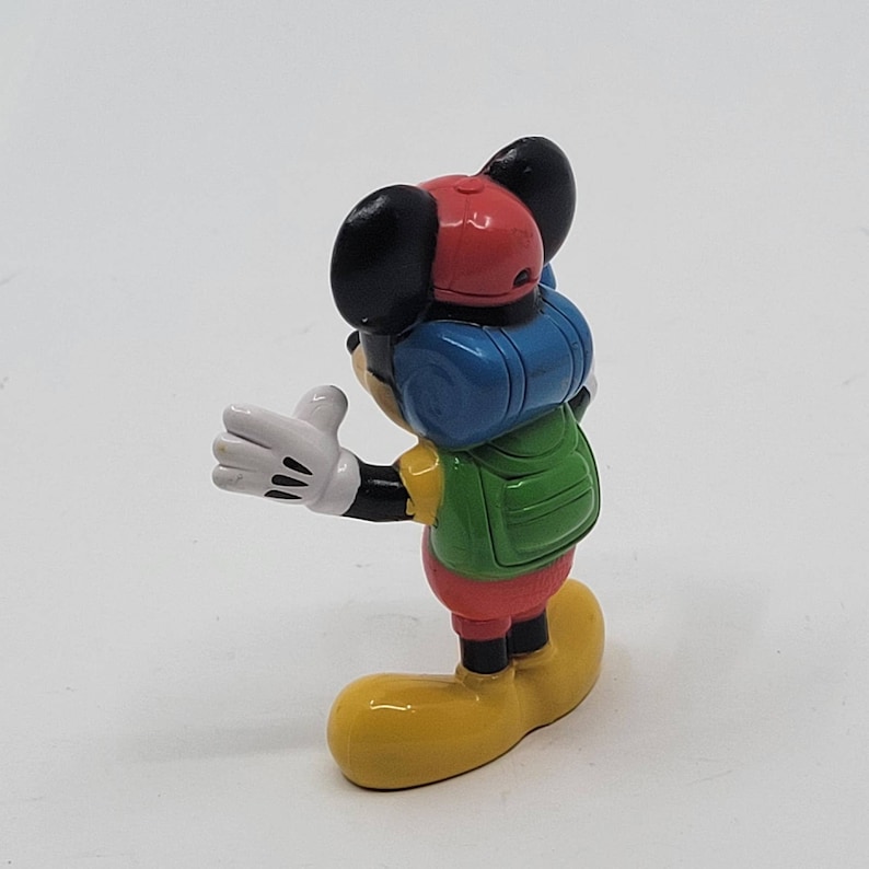 May include: A small plastic figurine of Mickey Mouse wearing a red hat, blue and green backpack, and yellow shoes.