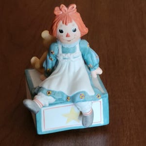 Può includere: Una statuetta in porcellana di Raggedy Ann seduta su una scatola di giocattoli blu e bianca con una stella gialla. Indossa un vestito blu con un grembiule bianco e ha un fiocco rosso nei capelli. Tiene un orsacchiotto.