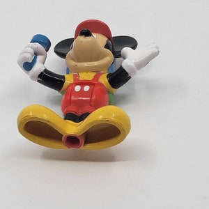 May include: A yellow, red and black plastic Mickey Mouse toy holding a blue microphone. The toy is standing with its legs spread apart.