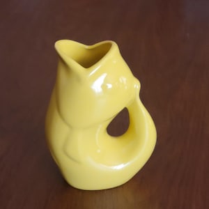 May include: A vibrant yellow ceramic jug with a heart-shaped spout and a distinctive, curved handle. The jug has a glossy finish and a smooth, organic form, suitable for serving drinks or as a decorative item.