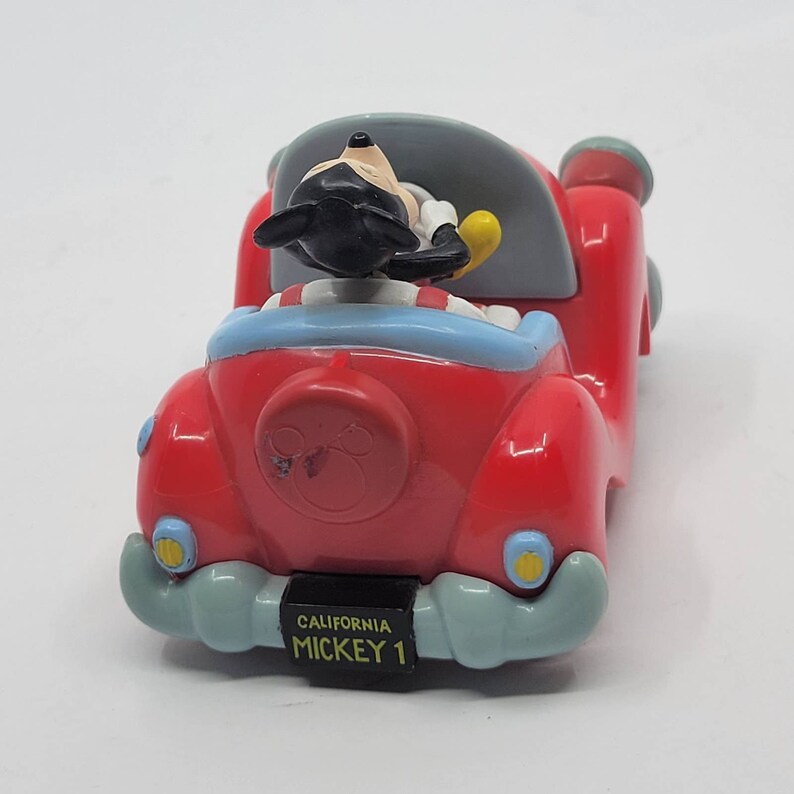 Puede incluir: Un coche de juguete rojo con una figura de Mickey Mouse al volante. El coche tiene un interior azul y blanco y un logotipo de cabeza de Mickey Mouse en la parte trasera. La matr&iacute;cula dice "California Mickey 1."