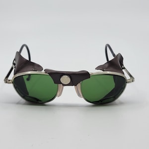 Puede incluir: Gafas de sol de estilo vintage con lentes verdes y montura plateada. Las gafas de sol tienen protectores laterales y puente de cuero marrón oscuro. Las patillas son negras y se curvan sobre las orejas. Las lentes son de color verde vibrante.