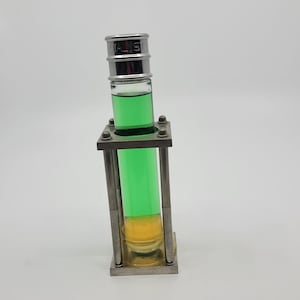 May include: A clear glass vial containing green and yellow liquids, held in a silver metal frame. The vial has a silver cap with the text "STALIS". The frame is rectangular with four vertical supports.