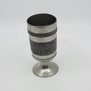 May include: A silver-colored goblet with intricate embossed designs. The cup features a detailed band of Celtic knotwork and a lower section with figures and swirling patterns. The goblet has a flared base and a cylindrical body, perfect for display.