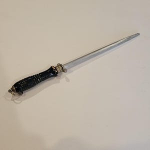 May include: A knife sharpening steel with a black handle and a silver sharpening rod. The handle has a decorative metal ring at the end. The sharpening rod is tapered to a point.