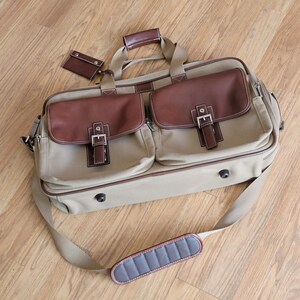 May include: A tan and brown briefcase with two front pockets, brown leather accents, and a matching luggage tag. The bag has a shoulder strap and a top handle. The bag is made of canvas and leather.