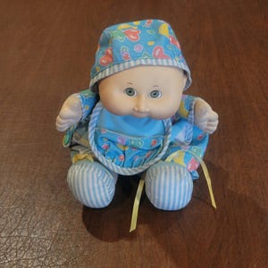 Puede incluir: Una muñeca de cuerpo blando con cara redonda, ojos verdes y tono de piel claro. La muñeca lleva un gorro azul y un conjunto a juego con un patrón colorido de corazones y formas, acentuado con tela de rayas azul claro y blanca y cintas amarillas.