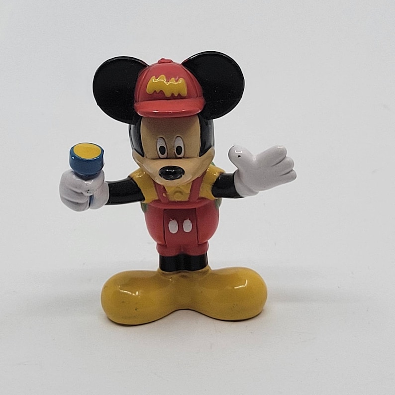 May include: A red and yellow Mickey Mouse figurine wearing a red hat with the letter "M" on it. He is holding a blue torch and has a white and green backpack.