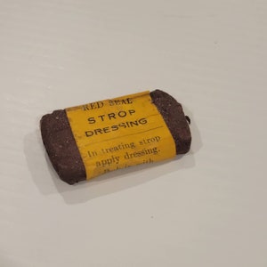 Puede incluir: Un pequeño bloque rectangular marrón de crema para strop con una etiqueta amarilla que dice "RED SEAL STROP DRESSING" y "In treating strop apply dressing."