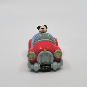 Puede incluir: Un coche de juguete rojo con una figura de Mickey Mouse al volante. El coche tiene un parabrisas azul, faros amarillos y una matr&iacute;cula que dice "California Mickey 1".