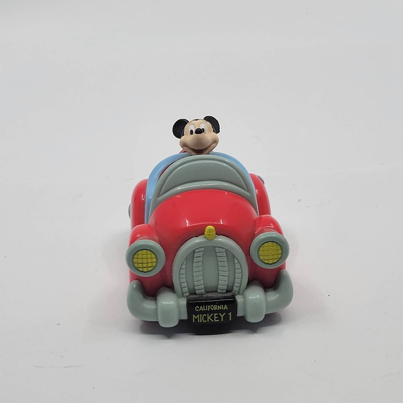 Puede incluir: Un coche de juguete rojo con una figura de Mickey Mouse al volante. El coche tiene un parabrisas azul, faros amarillos y una matr&iacute;cula que dice "California Mickey 1".