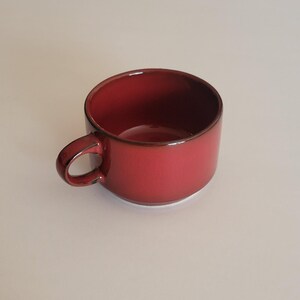 May include: A deep red ceramic mug with a smooth, glossy finish and a dark brown rim. The mug features a rounded handle on the side and a wide, open top. The interior of the mug is a matching deep red color, creating a consistent look throughout.