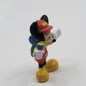 May include: A red, yellow and blue plastic figurine of Mickey Mouse wearing a red hat with a yellow logo, a green backpack and holding a blue torch. He is wearing red overalls with white polka dots and yellow shoes.