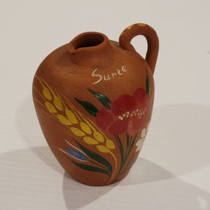 Puede incluir: Una pequeña jarra de terracota con asa, pintada con una flor roja, hojas verdes y trigo amarillo. La jarra tiene la palabra "Suerte" escrita en ella.