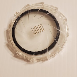 May include: Clear plastic camera lens filter with a black ring and the word "HOYA" in gold lettering. The filter has a ridged, circular design around the edge and a white interior.