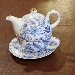 Może przedstawiać: Zestaw do herbaty dla jednej osoby z białej porcelany z niebieskim wzorem kwiatowym i złotym wykończeniem. Komplet zawiera dzbanek z pokrywką, filiżankę oraz pasujący spodek. Dzbanek stoi na filiżance, która spoczywa na spodku.