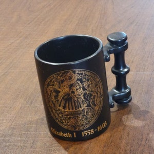 Pode incluir: Caneca de cerâmica preta com um design dourado do selo real da Rainha Elizabeth I e o texto "Elizabeth I 1558-1603".