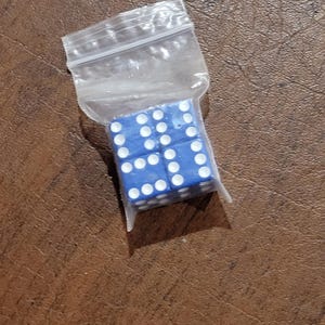 Puede incluir: Un conjunto de seis dados azules con puntos blancos, contenidos en una bolsa de plástico transparente. Los dados están dispuestos en forma cuadrada, mostrando cada uno un número diferente de puntos. La bolsa está parcialmente abierta, revelando los dados.