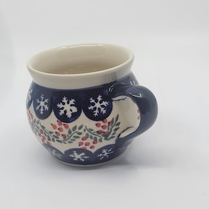 May include: A ceramic mug with a rounded shape and a dark blue handle. The mug has a white interior and is decorated with a pattern of snowflakes, red berries, and green leaves on a dark blue background.