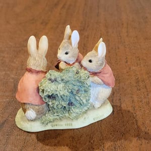 May include: A figurine featuring three small rabbits with light brown fur and white bellies, each wearing a pinkish-red jacket. The rabbits are positioned around a textured green bush on a light-colored base with the text FW & Co 1993 visible.