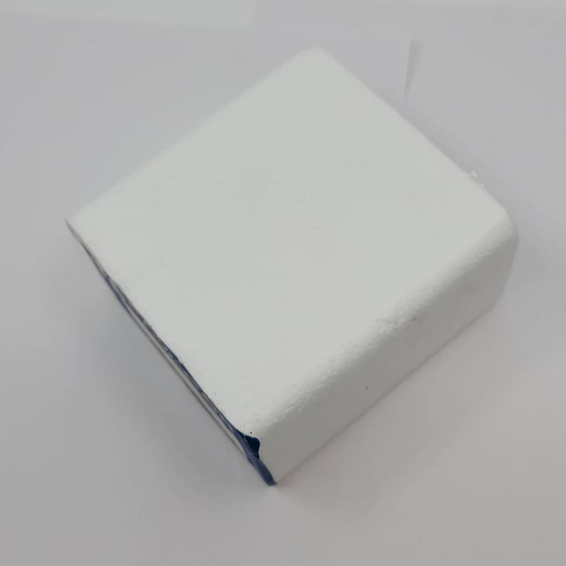 May include: A white, square-shaped object with a textured surface. The object has a rounded edge and a thin blue stripe along one side. The item is set against a white background.