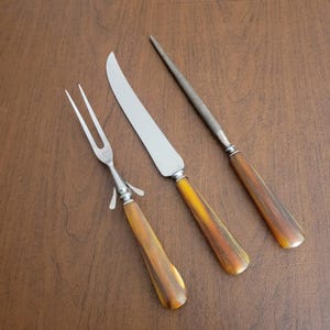 Könnte beinhalten: Ein dreiteiliges Tranchierbesteck bestehend aus einer zweizinkigen Fleischgabel, einem scharfen Tranchiermesser und einem Wetzstahl. Jedes Utensil hat einen polierten Metallkopf und einen glatten, konischen Griff mit braun-bernsteinfarbener Maserung.
