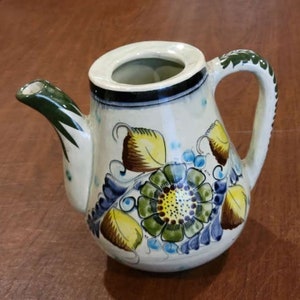 Könnte beinhalten: Eine weiße Teekanne aus Keramik mit einem grünen Henkel und Ausguss. Die Teekanne ist mit einem bunten Blumendesign verziert, das gelbe, grüne, blaue und braune Blumen und Blätter zeigt.