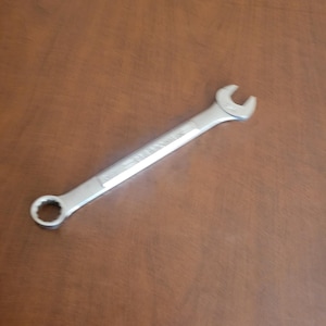 May include: A silver combination wrench, a hand tool with an open end and a box end. The wrench is lying on a brown surface. The tool is designed for tightening or loosening nuts and bolts.