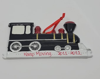 May include: Black and white train ornament with red accents and a red ribbon for hanging. The text "Keep Moving... 2011-2012" is written in red on the bottom. A decorative item for a Christmas tree or other holiday display.