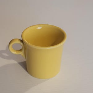 Puede incluir: Una taza de cerámica amarilla con un asa redondeada. La taza tiene forma cilíndrica y un acabado liso y brillante. El interior de la taza también es amarillo. La taza está sobre una superficie blanca.
