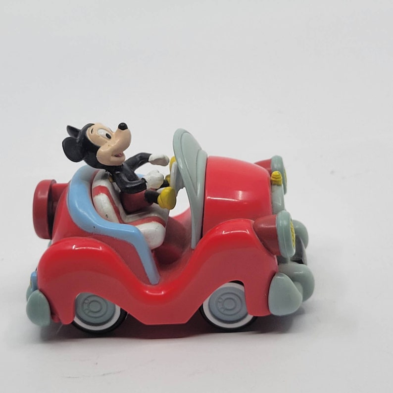 Puede incluir: Un coche de juguete rojo con una figura de Mickey Mouse al volante. El coche tiene un asiento a rayas azul y blanco y detalles grises.