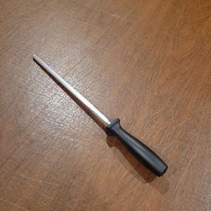 Puede incluir: Una taza de cerámica blanca con asa negra y el texto "But First, Coffee" en negrita negra. La taza se encuentra sobre un fondo borroso, sugiriendo un ambiente acogedor.