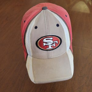 Peut inclure: Casquette de baseball rouge, beige et blanche avec le logo des 49ers de San Francisco. La casquette a une visière incurvée et des panneaux latéraux en mesh. Le logo de l'équipe est brodé sur le devant.