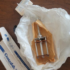 May include: An electronic component, possibly a potentiometer or switch, is partially unwrapped in tissue paper. The component has a metal shaft and multiple terminals. Packaging includes a box with the text "Centralab" and "Milwaukee, Wisconsin."