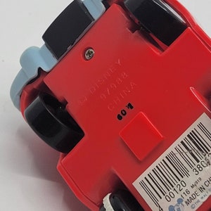 Puede incluir: Coche de juguete rojo con ruedas negras y techo azul y blanco. El coche tiene un logotipo de Disney y el texto "97988 CHINA 601" impreso en la parte inferior. El coche tambi&eacute;n tiene un c&oacute;digo de barras y el texto "00120 M919 38032 MADE IN CH".