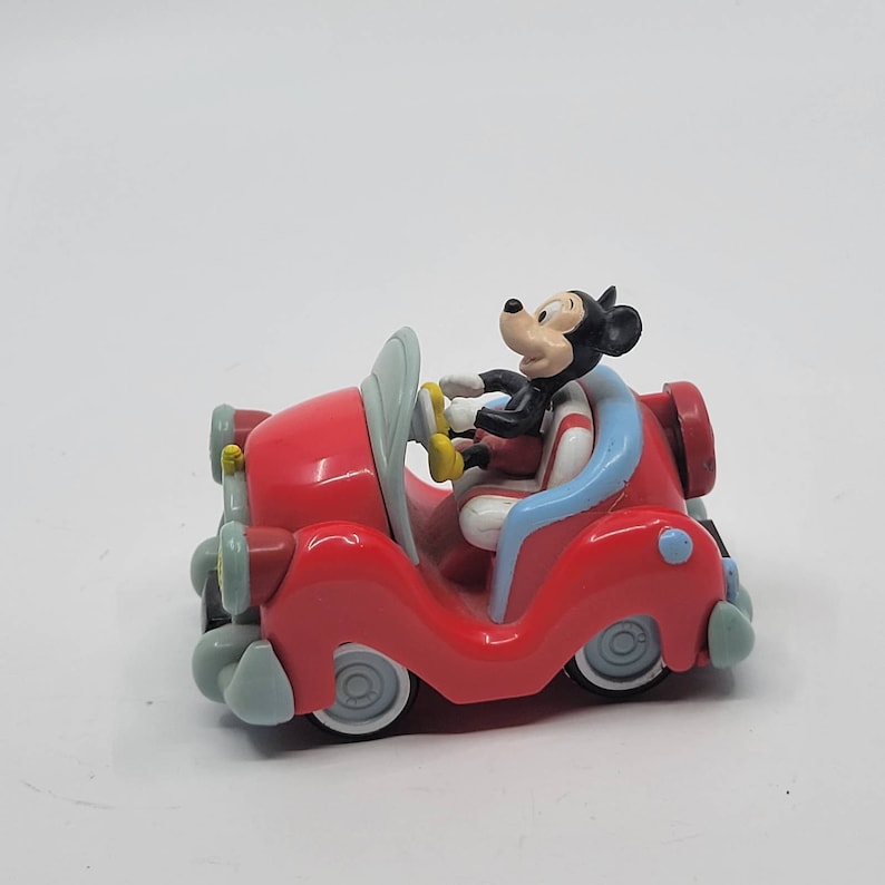 Puede incluir: Un coche de juguete rojo con un interior azul y una figura de Mickey Mouse al volante. El coche tiene un volante blanco y neum&aacute;ticos negros.