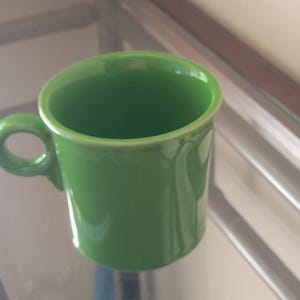 May include: A solid lime green ceramic mug with a smooth, glossy finish. The mug features a cylindrical body with a slightly flared rim and a small, rounded handle attached to the side. The interior and exterior are a uniform, vibrant green colour.