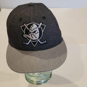 May include: A dark grey baseball cap with a grey brim. The cap features a white and black embroidered logo of a hockey mask and crossed hockey sticks. The cap is sitting on a glass object.