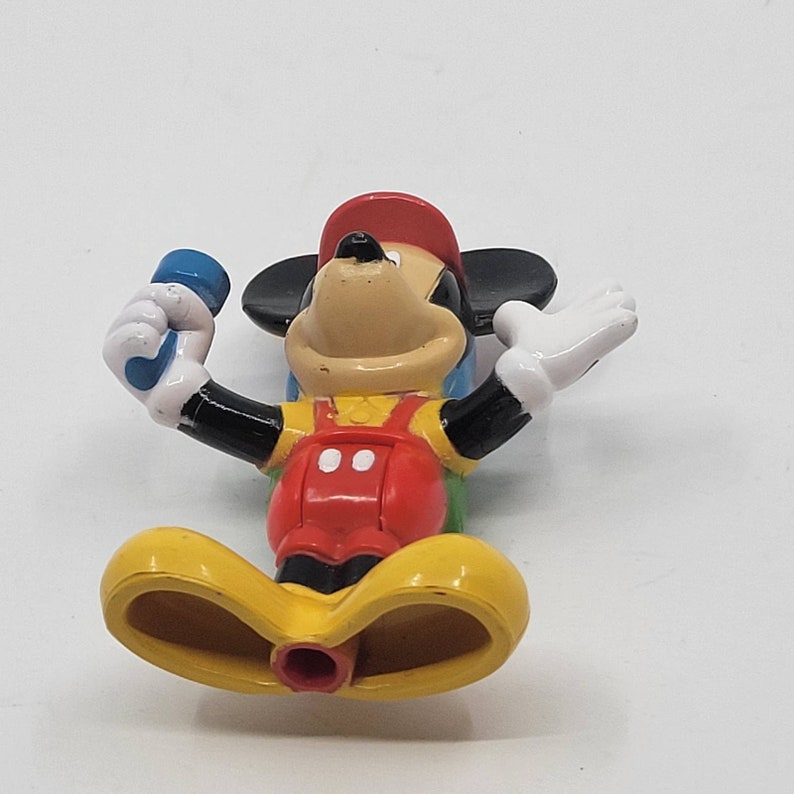 May include: A red, yellow and blue plastic Mickey Mouse toy with a blue microphone. The toy is wearing a red hat and overalls.