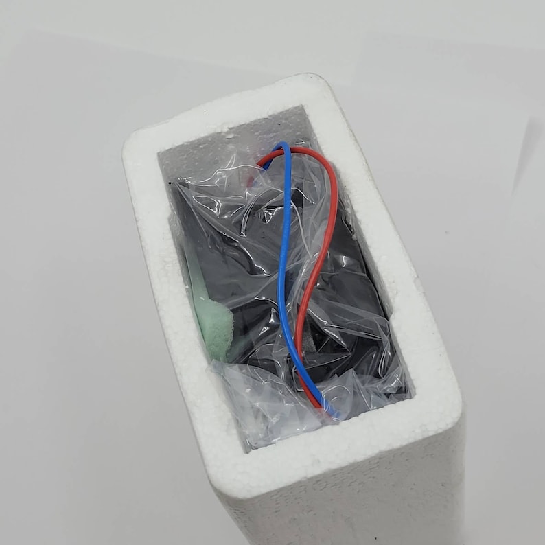 May include: A black electronic component, wrapped in clear plastic, is nestled inside a white foam container. Red and blue wires extend from the component. The foam packaging is rectangular and has a smooth texture.