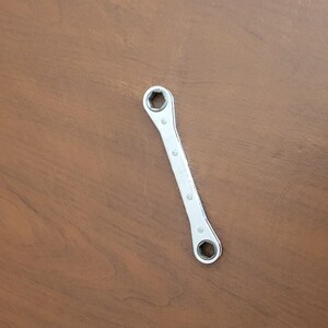 Op de afbeelding: Een zilveren, gebogen sleutel met zeshoekige openingen aan elk uiteinde. De tool heeft een reeks kleine klinknagels over de lengte en ligt op een bruin houten oppervlak. Deze handtool is ontworpen voor het aandraaien of losdraaien van moeren en bouten.
