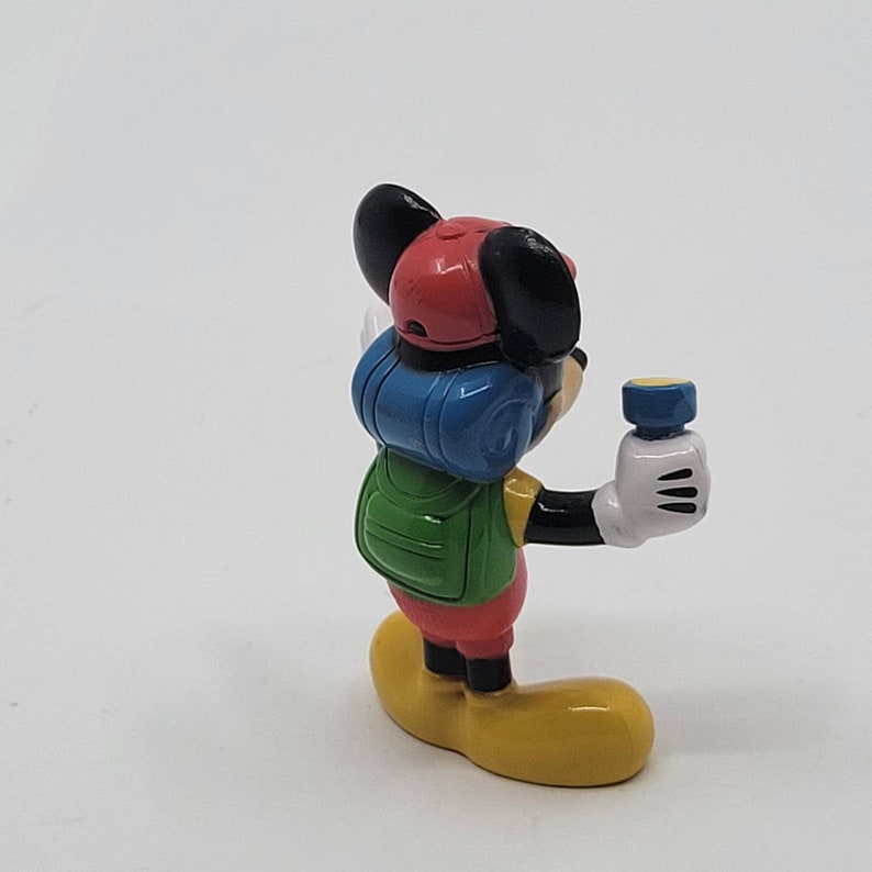May include: A small plastic figurine of Mickey Mouse wearing a red hat, blue and green backpack, and holding a torch.
