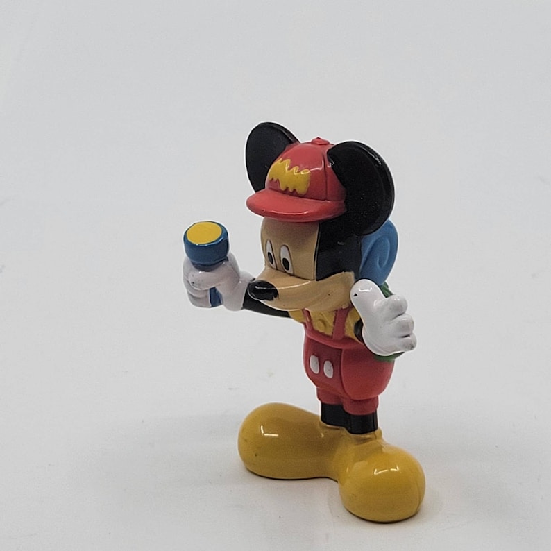 May include: A red, yellow and blue plastic figurine of Mickey Mouse wearing a red and yellow hat and holding a blue torch. The figurine is standing on a white surface.